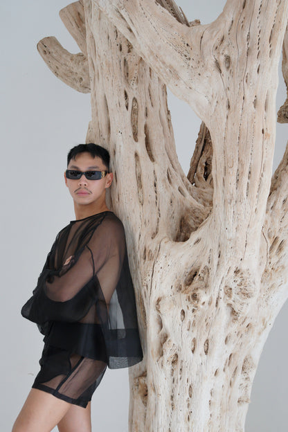 Person wearing a black outfit in sheer Silk Organza set standing under a large piece of driftwood.